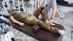 Photo: a young man in camouflage pants and dark green T-shirt lies on a physiotherapy couch and is being treated by a man; Copyright: envato/Iakobchuk 