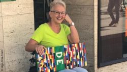Photo: a woman with light grey hair, glasses and a green t-shirt is sinnting in a wheelchair in front of a building, on her legs she carries a ramp made of tiny bricks; Copyright: Lego-Oma-Team