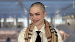Nika Sky smiles directly at the camera. She is wearing a white shirt with a tie and a brown scarf draped over her shoulders.