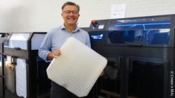 Jacob M. Nissen, CEO of Create it REAL, stands in front of industrial 3D-printing equipment holding a large white lattice-structured wheelchair cushion made with Programmable Foam®.