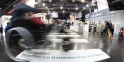 Photo: wheelchair user in a blurred photo effect drives over a people with disabilities and jobs sign at REHACARE trade fair; Copyright: Messe Düsseldorf/ctillmann